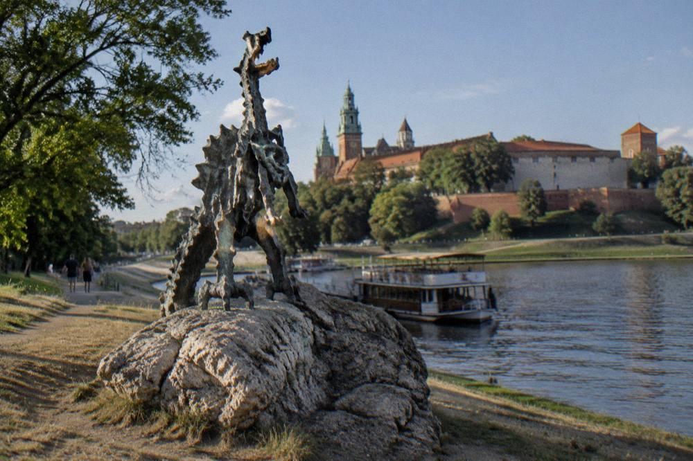 Bronze dragon statue on a rocky Vistula embankment below Wawel Hill, with a moored boat on the river.