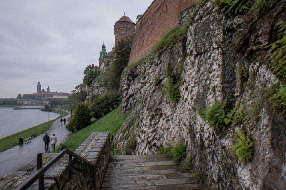Wet stone steps run below Wawel Hill limestone cliffs and brick fortifications beside the Vistula river path.