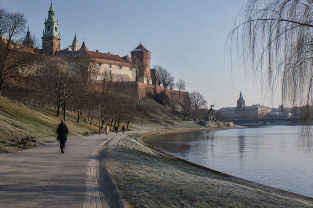Frosty Vistula riverside path curves below Wawel Hill, with bare trees, distant walkers and the dragon statue near the bank.