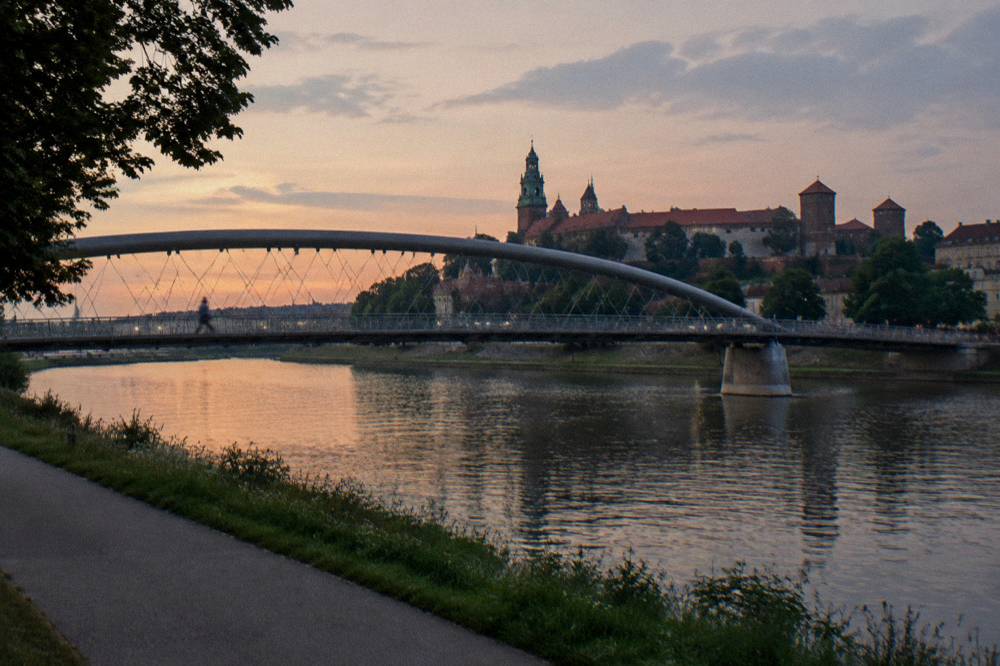 Curved footbridge over the Vistula at dusk with Wawel Castle on the hill behind and a riverside path in the foreground.